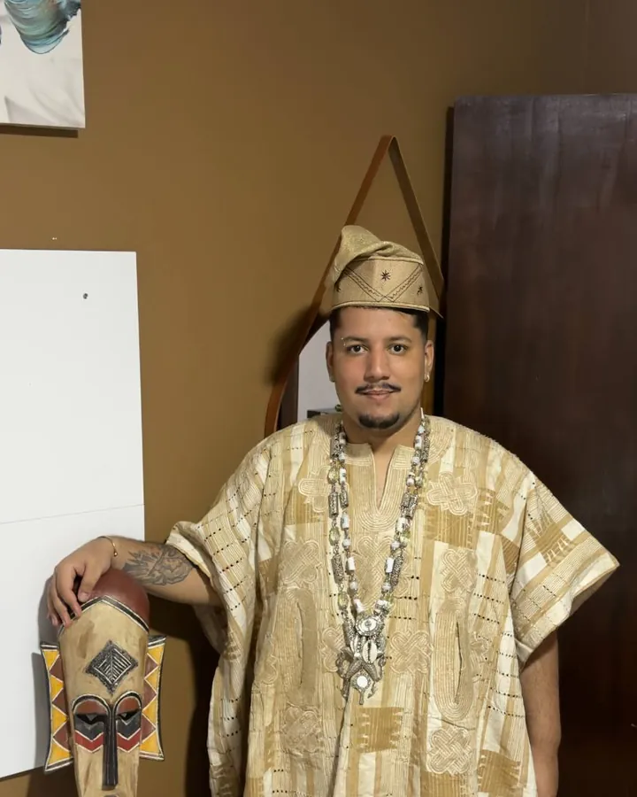 Pai de Axé de pé, com roupa tradicional africana bordada em dourado, chapéu e colares de contas, ao lado de uma máscara africana de madeira